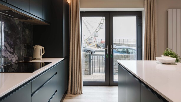 A modern kitchen with dark cabinets, a black countertop, and a view of the street through a glass door. - Experience a sense of deep calm with seaside walks & blissful sea air (Portrush)