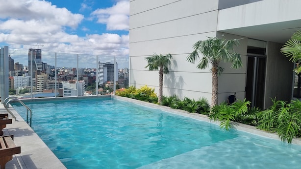 Una piscina en la azotea con agua cristalina azul, rodeada de palmeras y plantas, con vistas a un paisaje urbano con edificios modernos.