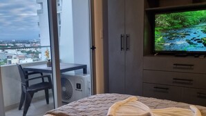 Un dormitorio con cama, televisión, balcón con una silla y vistas a la ciudad.