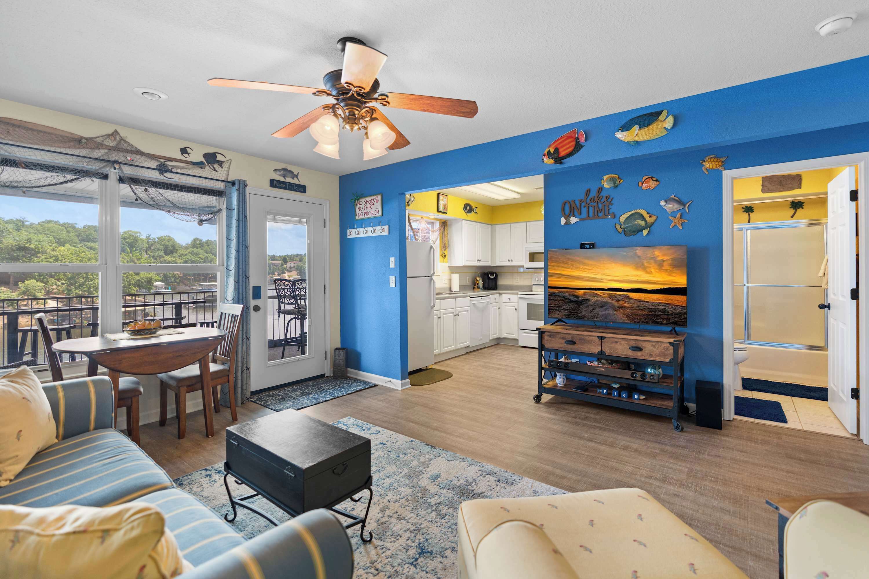 A spacious living area with a large window, a ceiling fan, a dining table with chairs, a sofa, a coffee table, and a kitchen area with cabinets and a sink.