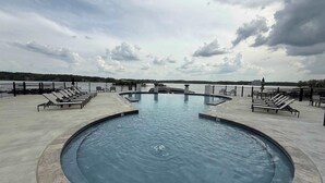 Una piscina circular con sillones de descanso alrededor, una estructura de piedra en el centro y vista a un cuerpo de agua y colinas al fondo.