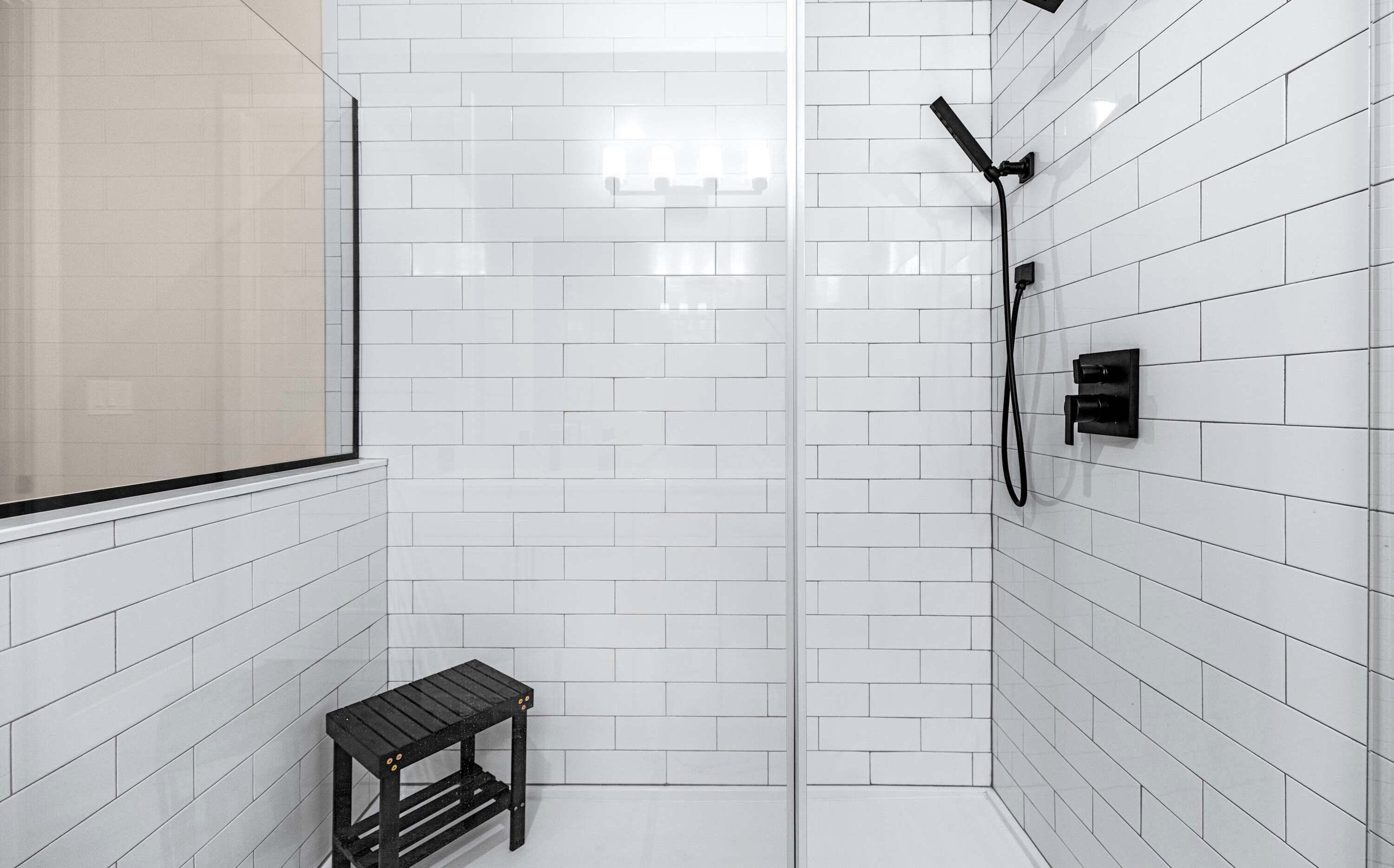 Une salle de bain moderne avec des carreaux de métro blancs, une cabine de douche en verre, une douchette noire et un petit tabouret en bois.
