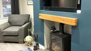 Ein gemütliches Wohnzimmer mit einem grauen Sofa, einem Holzregal, einem schwarzen Kaminofen und einem an der Wand montierten Fernseher.
