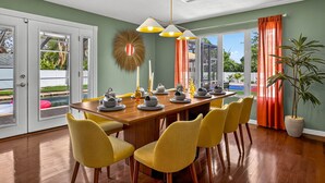 Un coin repas avec une table en bois dressée pour quatre personnes, des chaises jaunes et une grande fenêtre ornée de rideaux orange.