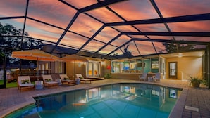 Une piscine entourée de chaises longues et de parasols, avec une terrasse couverte offrant une vue sur le coucher du soleil.