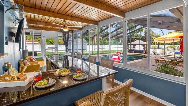 Une cuisine moderne dotée d’un grand îlot central, d’un plan de travail en verre et offrant une vue sur un jardin avec une piscine et du mobilier d’extérieur.