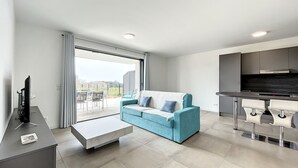 A modern living room with a sofa, a coffee table, and a TV. There is a sliding glass door leading to an outdoor area with a table and chairs.