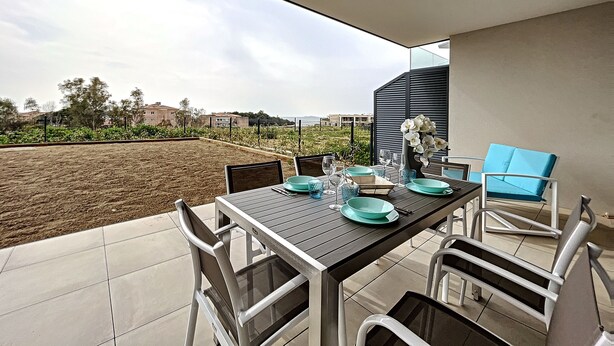 A dining area with a table set for four, outdoor chairs, and a view of a garden and buildings in the background.