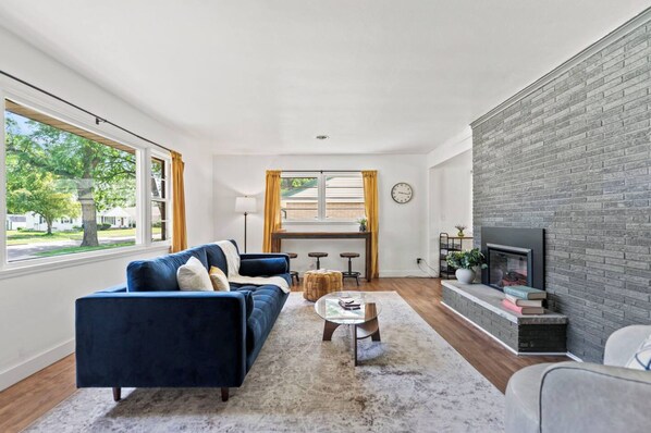 A living room with a blue sofa, a fireplace, and a view of the outdoors. - Entertainment Haven! Firepit, Game Room, and Large fenced-in yard (Janesville)