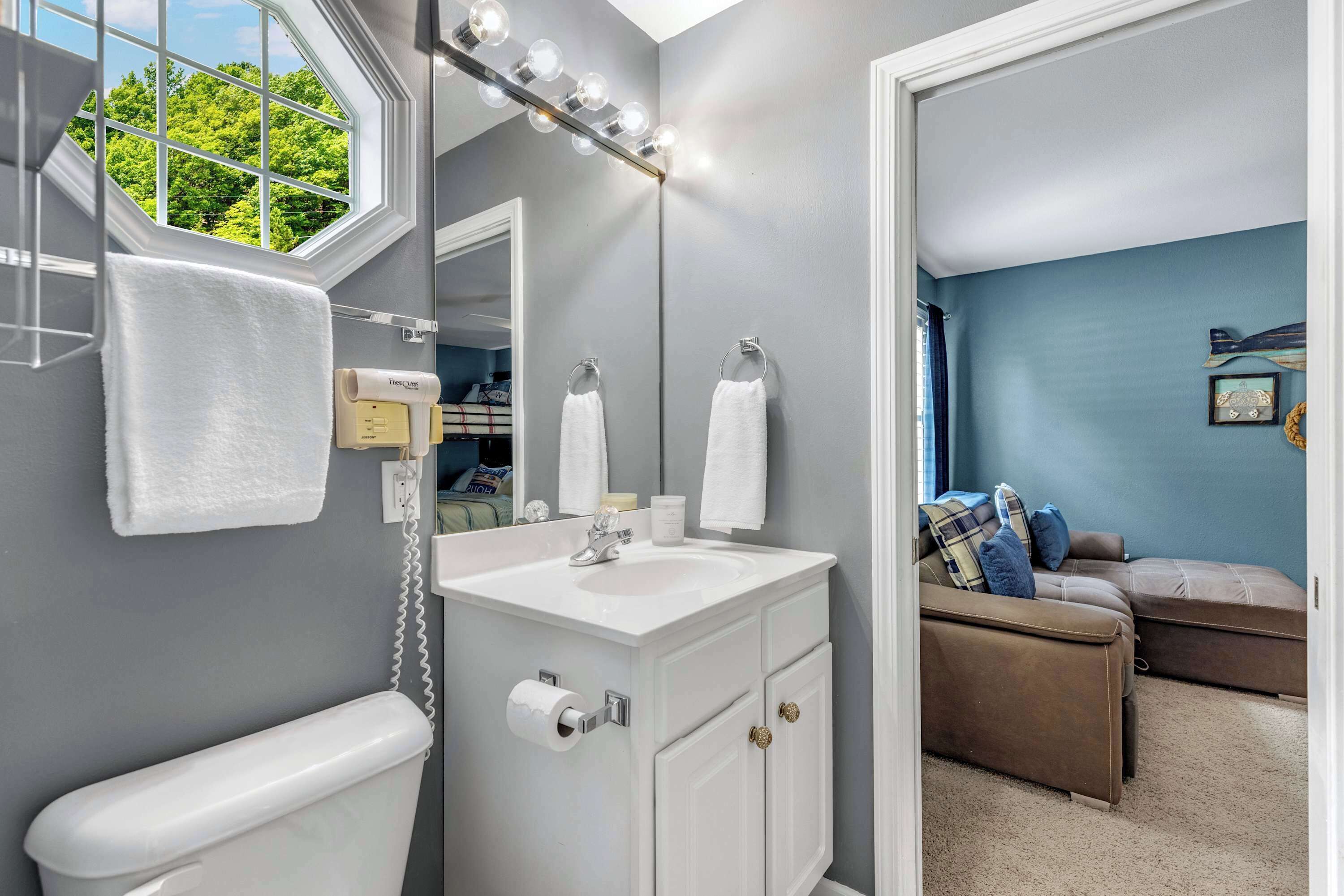 A bathroom with a toilet, sink, and vanity, a window with a view of trees, and a mirror with lights.