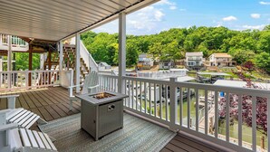 A covered deck with a grill, white Adirondack chairs, and a view of a residential area with houses and a dock. - Shore Thing - Lakefront for 12 (Lake Ozark)