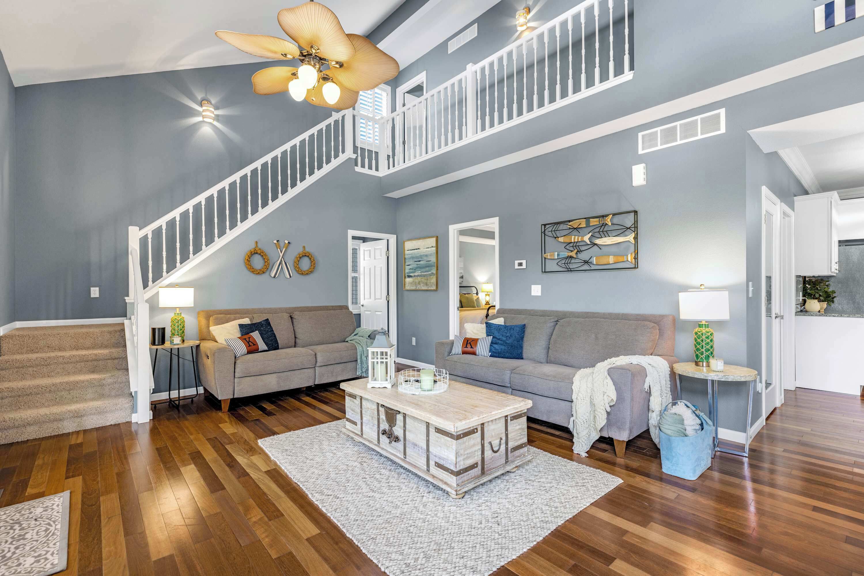 A spacious living room with hardwood flooring, a staircase with white railings, a large ceiling fan, and a cozy seating area with a sofa and armchairs.