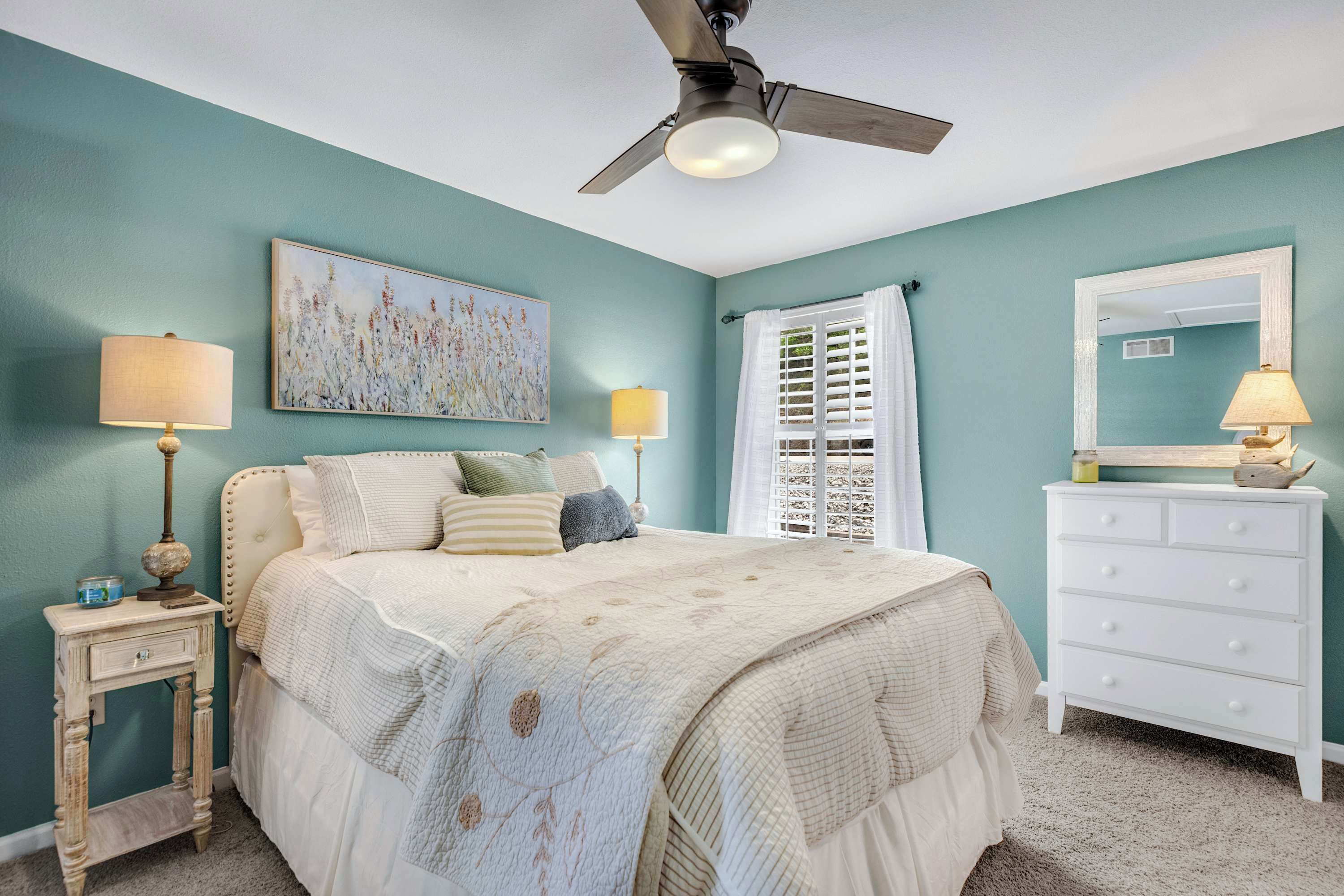 Quarto com paredes verde-água, cama com cabeceira branca, ventilador de teto, mesa de cabeceira com luminárias e cômoda.