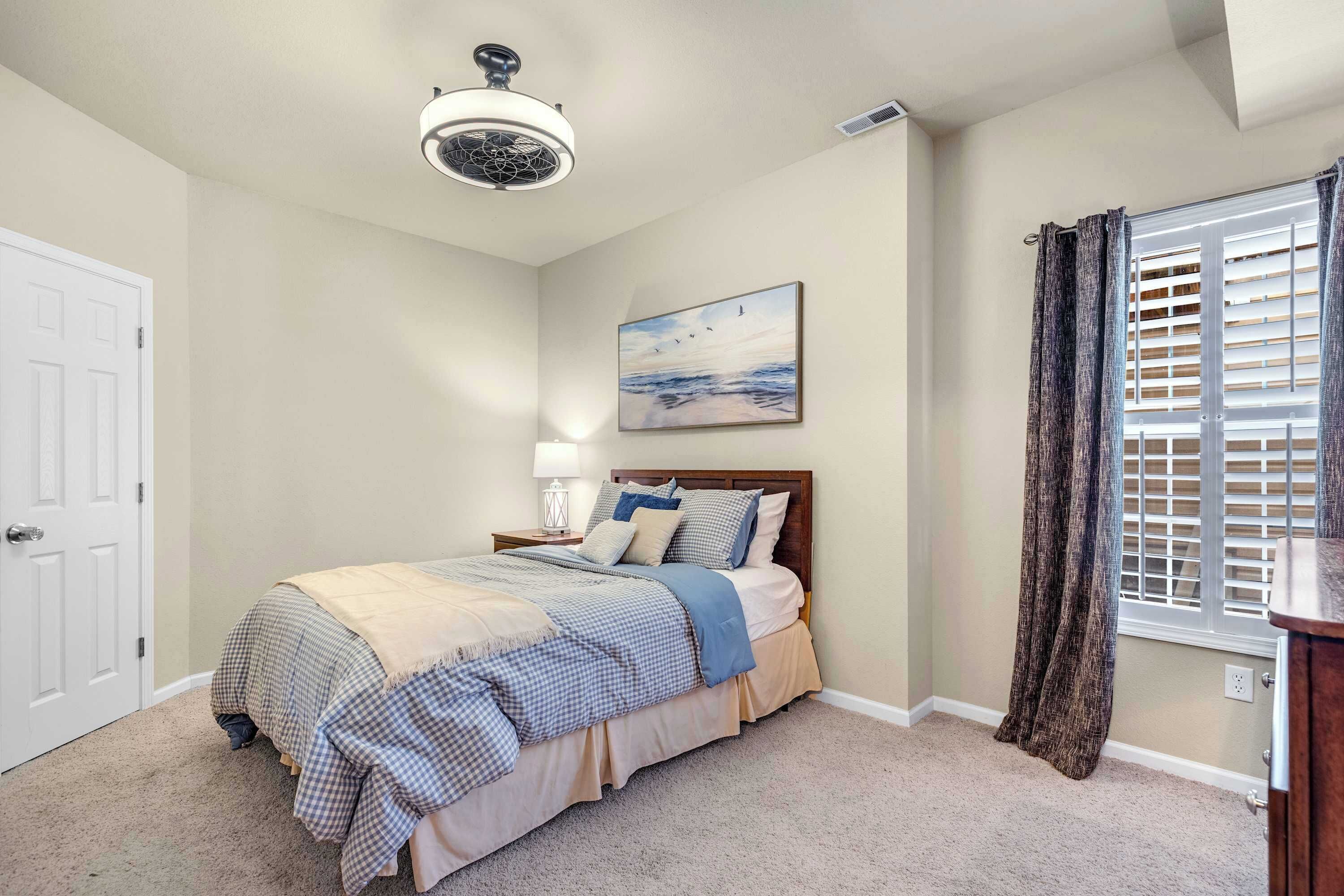 Un dormitorio con cama, ventilador de techo, ventana con persianas y un cuadro en la pared.