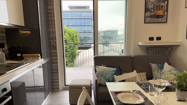 Une cuisine moderne avec un coin repas, un canapé et un balcon offrant une vue sur les bâtiments et la verdure.