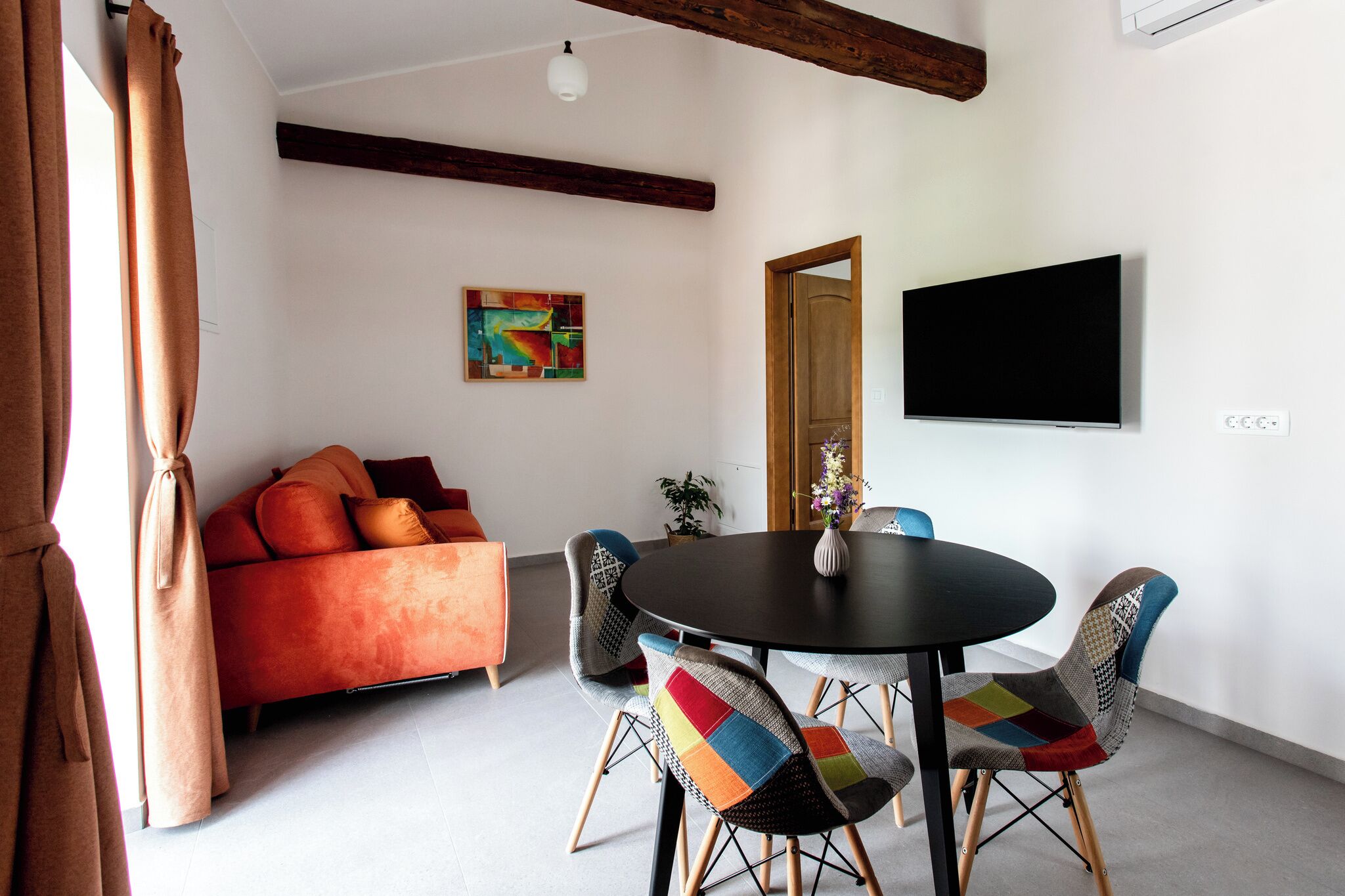 A modern living room with a sofa, a round table, and two chairs with colorful cushions. A flat-screen TV is mounted on the wall, and a painting is visible on the wall.