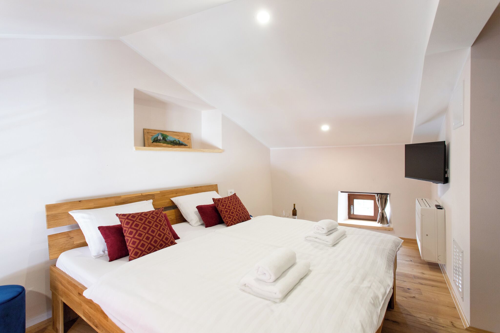 Quarto com cama de madeira, roupa de cama branca, TV na parede e uma pequena janela com cortinas.