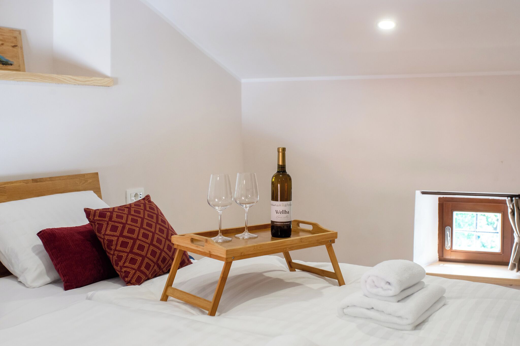 A hotel room with a bed, a wooden tray with a bottle of wine and two glasses, a window with curtains, and a red patterned pillow.