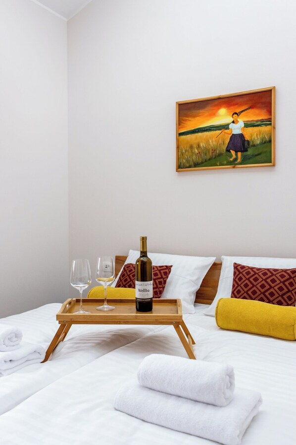 A hotel room with a bed, a painting of a woman in a field, a tray with a bottle of wine, glasses, and towels.