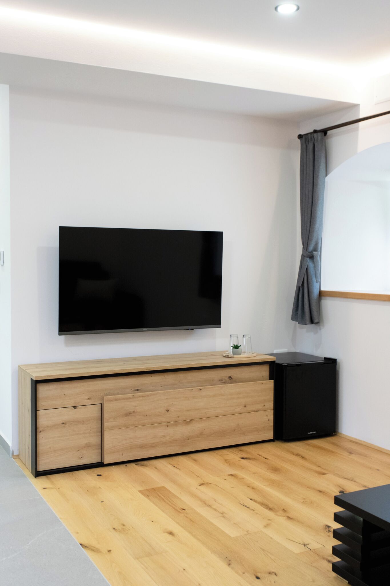 A modern living room with a flat-screen TV mounted on a wooden cabinet, a grey curtain, and a black speaker.