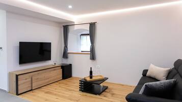 A modern living room with a flat-screen TV, wooden flooring, a black sofa, and a small table with a bottle and glasses.