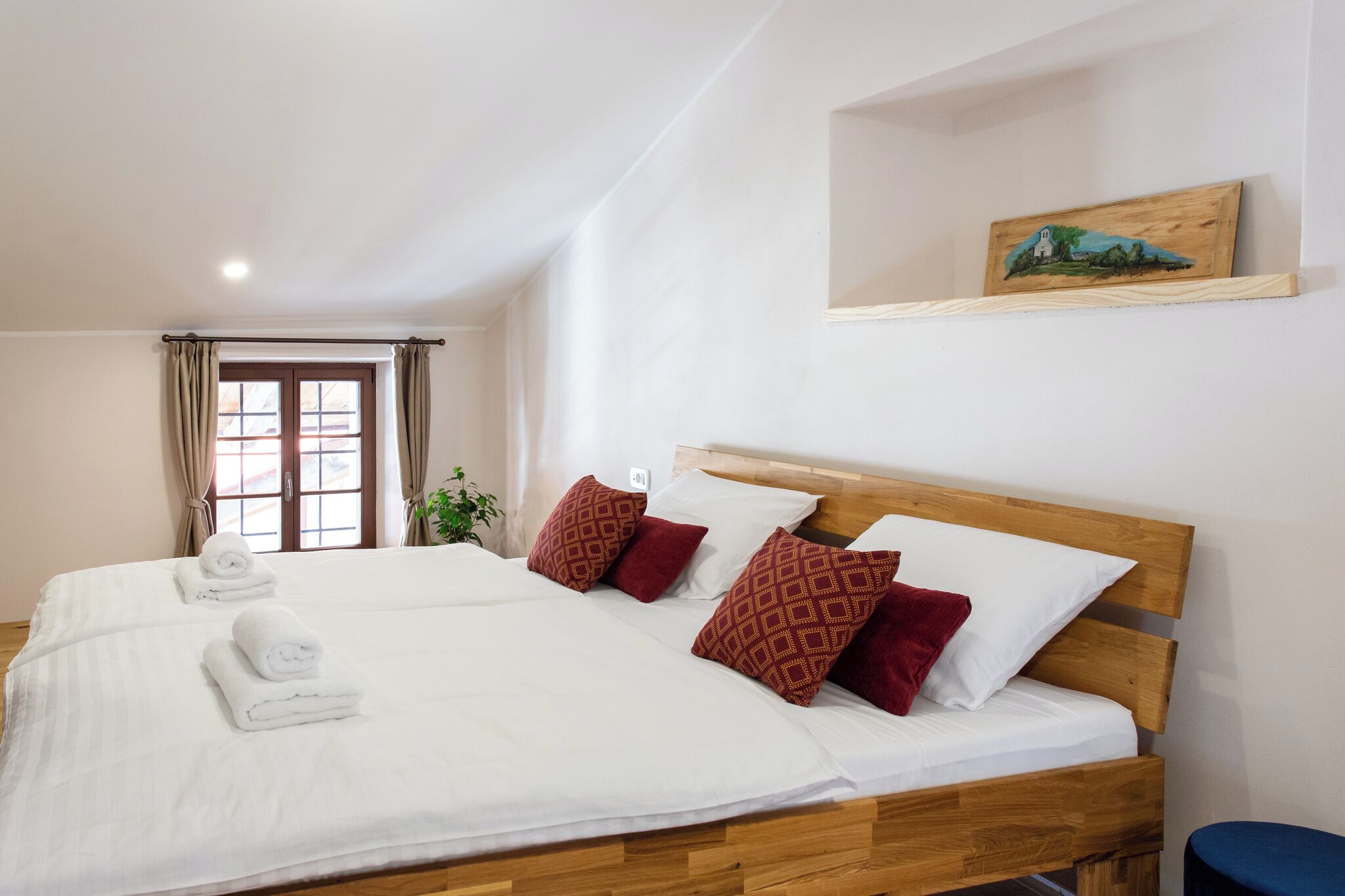 A double bed with white linens and red patterned pillows, a wooden headboard, and a small shelf with a painting.