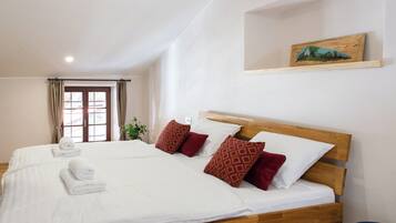 A double bed with white linens and red patterned pillows, a wooden headboard, and a small shelf with a painting.