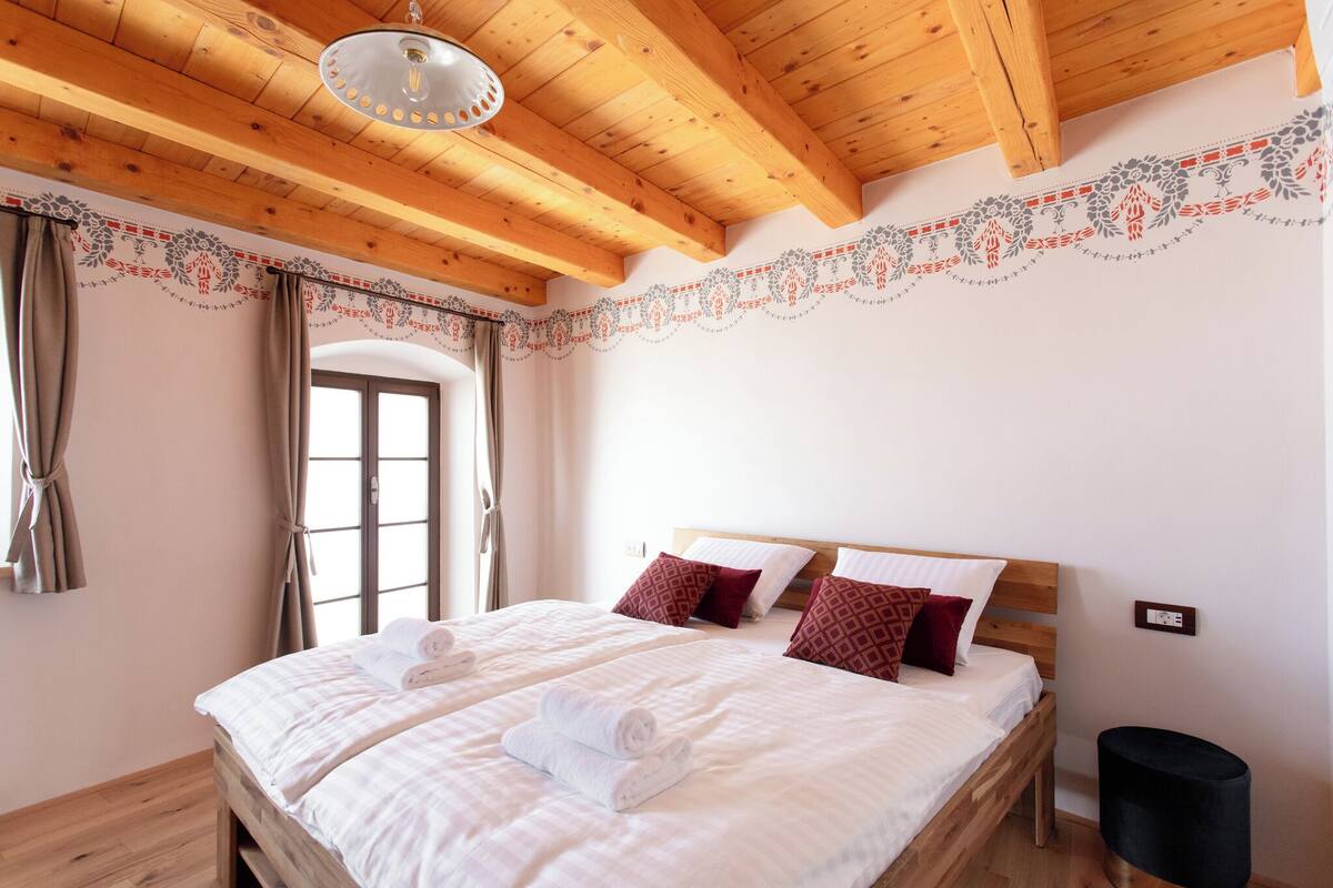 Uma cama de casal com lençóis brancos e almofadas vermelhas estampadas, teto de madeira e uma moldura decorativa na parede.
