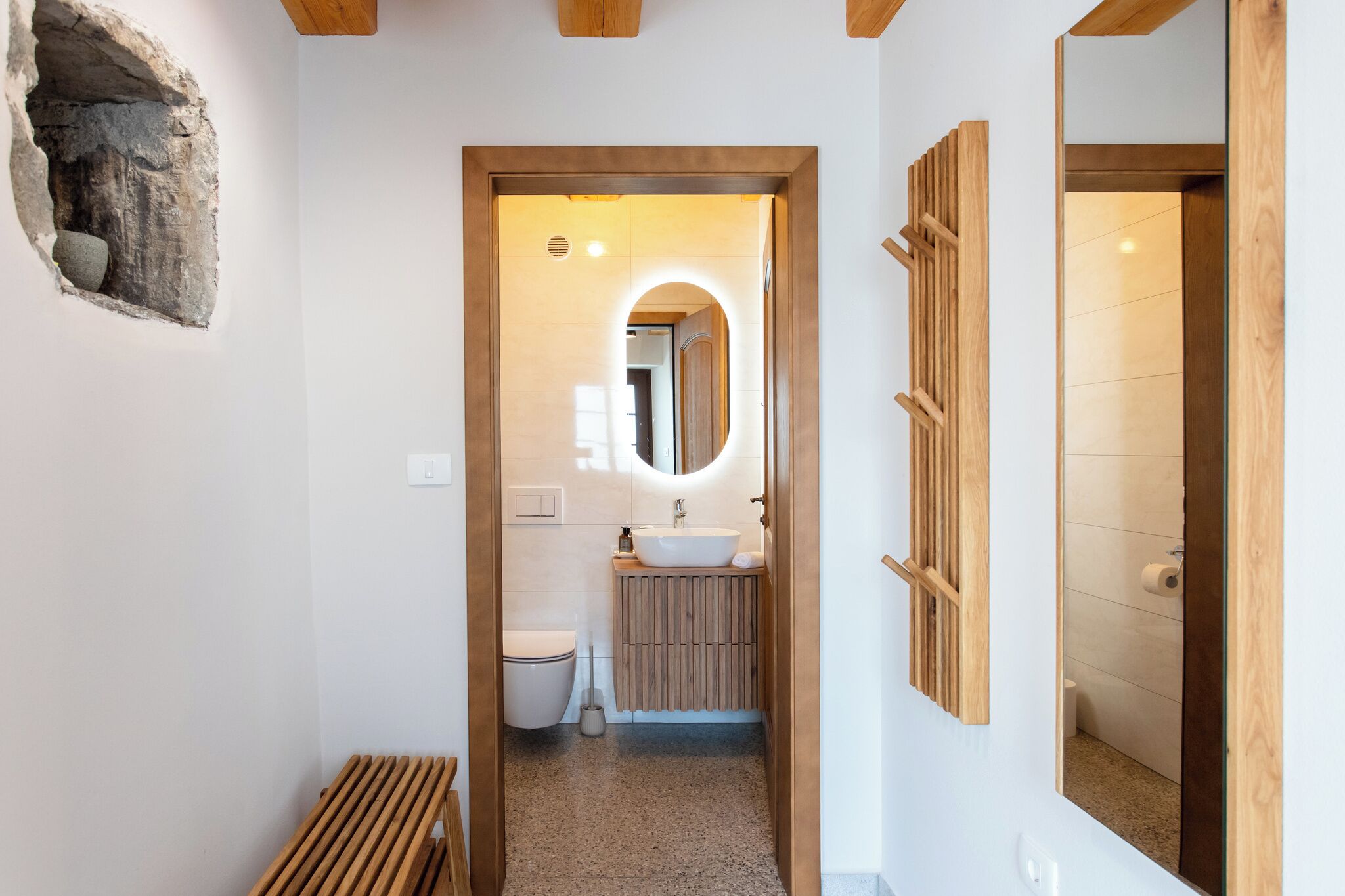 Een smalle gang met een houten bankje, een wanddecoratie van steen, een badkamer met een houten wastafelmeubel, een toilet en een spiegel.