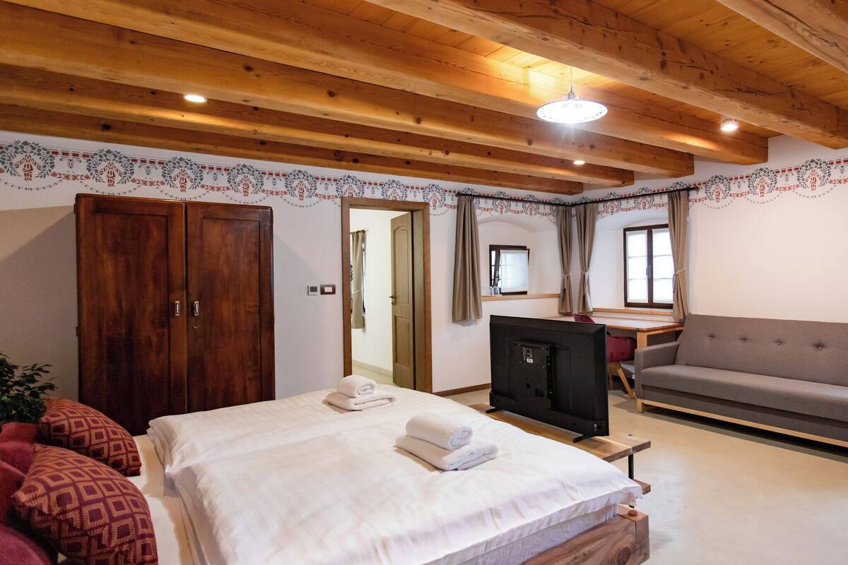 Quarto com teto de madeira, cama com roupa de cama branca, sofá cinza, televisão e guarda-roupa de madeira.