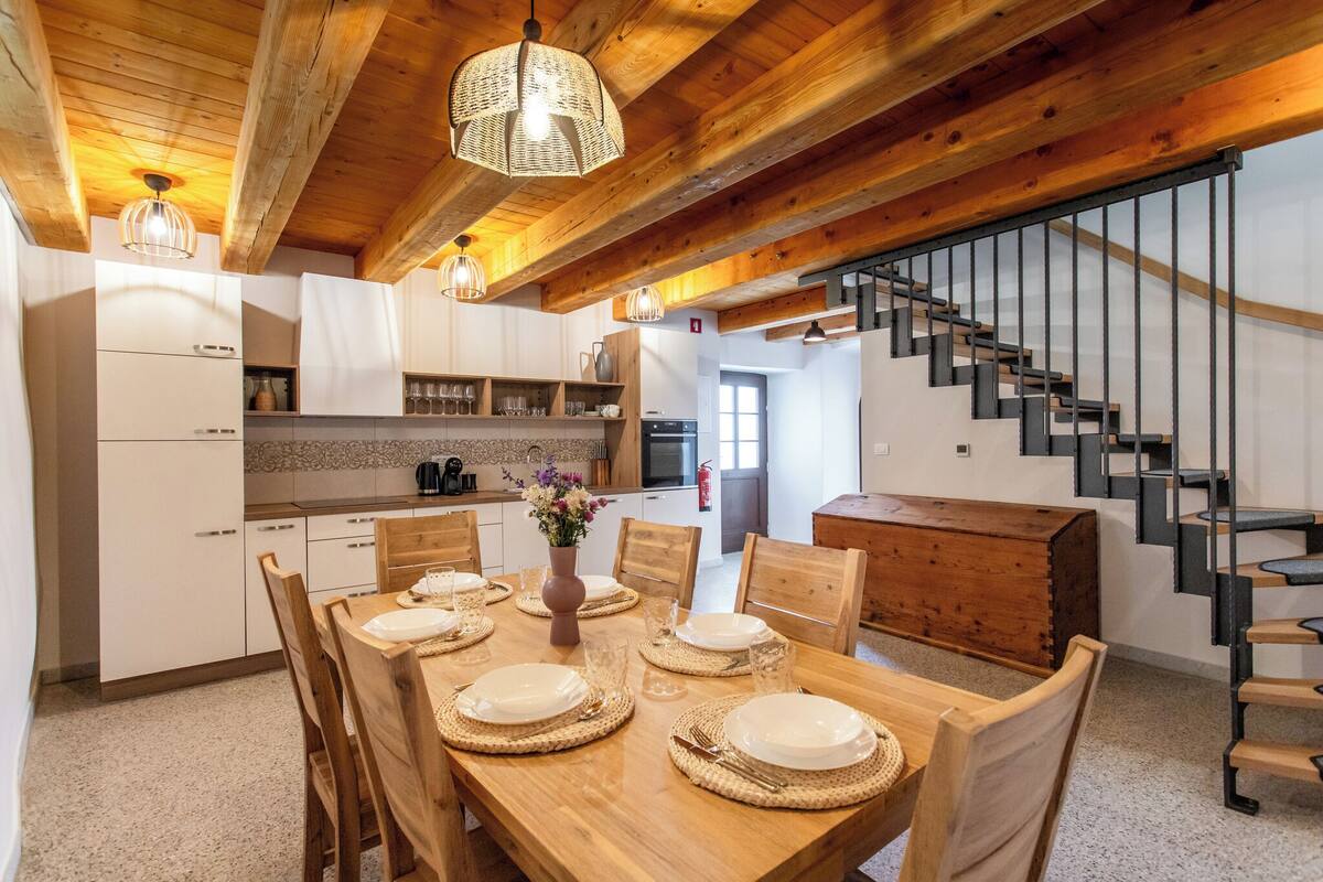 Uma cozinha moderna com armários brancos, uma mesa de jantar de madeira preparada para quatro pessoas e uma escada com corrimão preto.