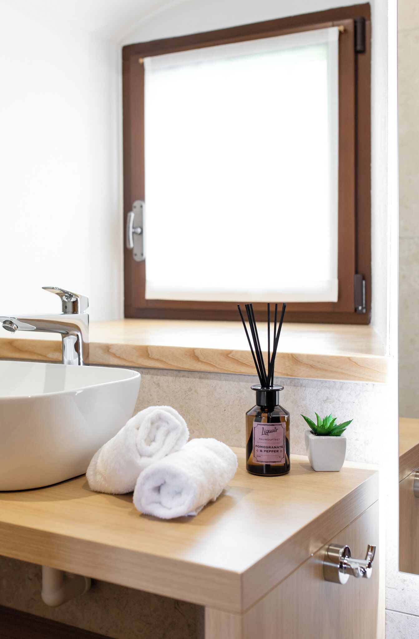 A bathroom with a large white sink, a mirror, a window with a brown frame, a bottle of reed diffuser, and a small potted plant.