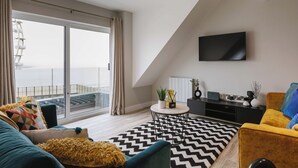 A modern living room with a large sliding glass door leading to a balcony, a flat-screen TV mounted on the wall, and a black and white chevron-patterned rug. - VISIT THE LOOKOUT AND EXPERIENCE THE ATLANTIC OCEAN AT ITS BEST. (Portrush)