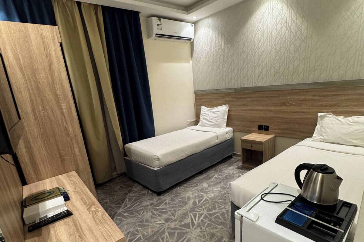 A hotel room with a bed, a desk with a kettle, a small table, and an air conditioning unit.