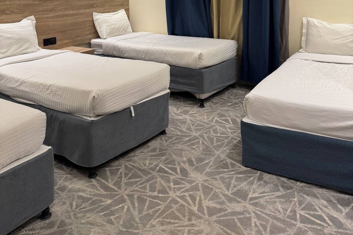 A hotel room with three beds, a wooden headboard, patterned carpet, and a window with blue and beige curtains.