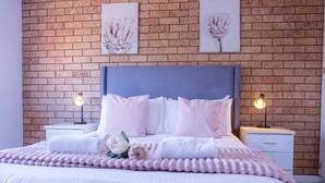 Una cama impecable con cabecera azul, ropa de cama blanca y toques de color rosa. Hay dos mesitas de noche con lámparas, y la pared muestra un diseño de ladrillo con dos obras de arte enmarcadas.