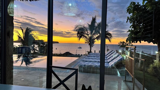 A view from a balcony overlooking a beach with a sunset, palm trees, and a small boat in the distance. - Oceanfront Luxury Villa with Infinity Pool and Private Beach (Nosy Be)