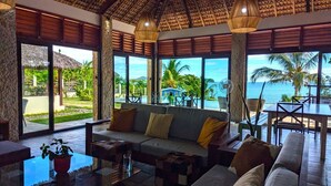 A spacious living area with a large sofa, a coffee table, and a dining table. The room features large windows offering a view of the ocean and a tropical landscape. - Oceanfront Luxury Villa with Infinity Pool and Private Beach (Nosy Be)