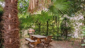 A wooden picnic table and benches in a garden with dense foliage and a palm tree.
