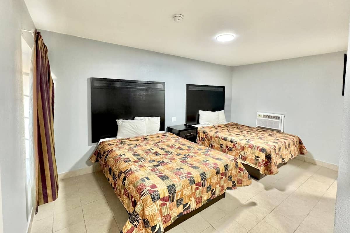 A hotel room with two beds, each with a patterned bedspread, a nightstand, a TV, and a wall-mounted air conditioning unit.