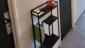 Un pasillo con una puerta negra, un espejo montado en la pared y una estantería negra con libros y un jarrón.
