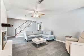 A modern living room with a sectional sofa, a coffee table, a ceiling fan, and a staircase.