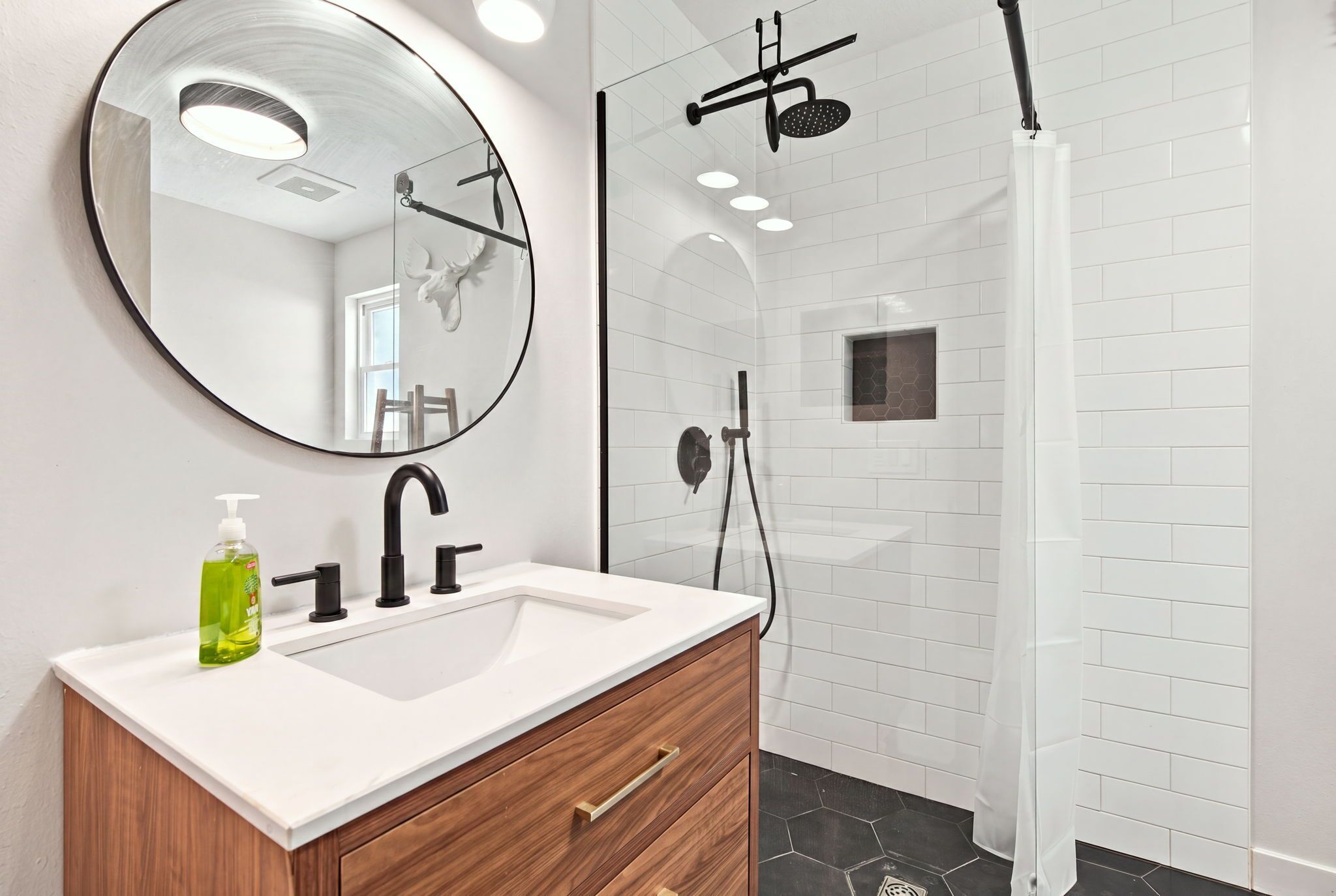 A bathroom with a white vanity, a round mirror, a shower with a black hose, and a hexagonal tile pattern on the walls.