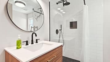 A bathroom with a white vanity, a round mirror, a shower with a black hose, and a hexagonal tile pattern on the walls.