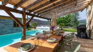 Una pérgola de madera con mesa de billar instalada debajo, con vista a una piscina y un área de barbacoa.