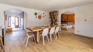 Un amplio espacio habitable con una mesa de comedor de madera preparada para cuatro personas, rodeada de sillas blancas. Se observa una escalera de madera y un sofá al fondo.