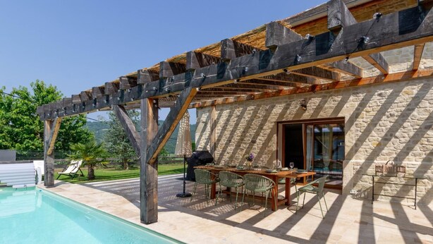 Zona junto a la piscina con un pérgola, pared de piedra y mobiliario exterior.