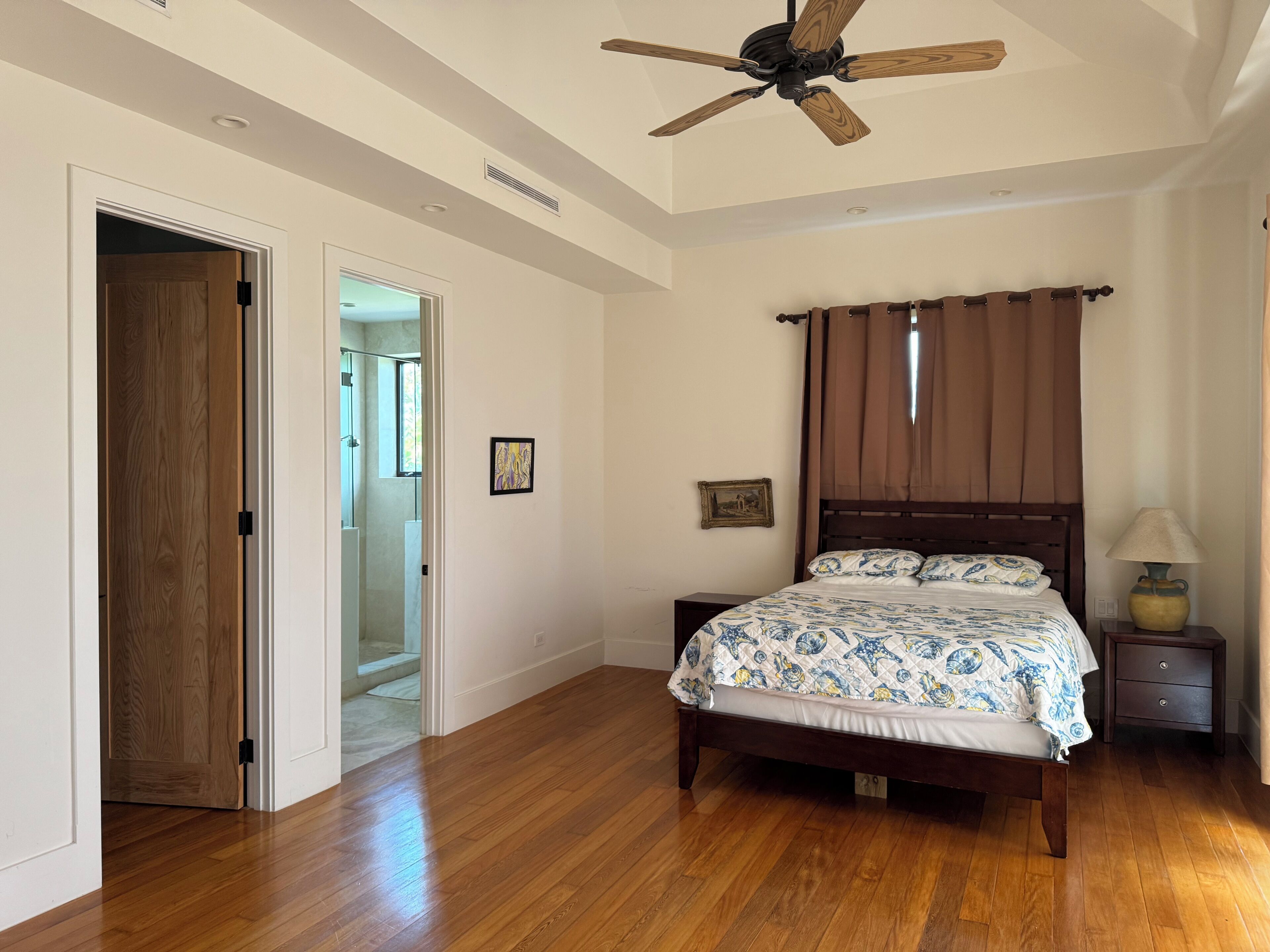 Une chambre avec un plancher en bois, un lit recouvert d’une couvre-lit à motifs, un ventilateur de plafond et une table de chevet avec une lampe.
