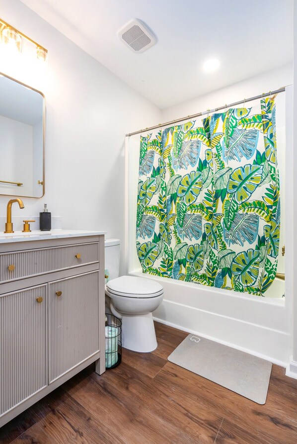 Une salle de bain avec un meuble-lavabo blanc, un robinet doré, des toilettes et un rideau de douche orné d’un motif de feuilles tropicales.