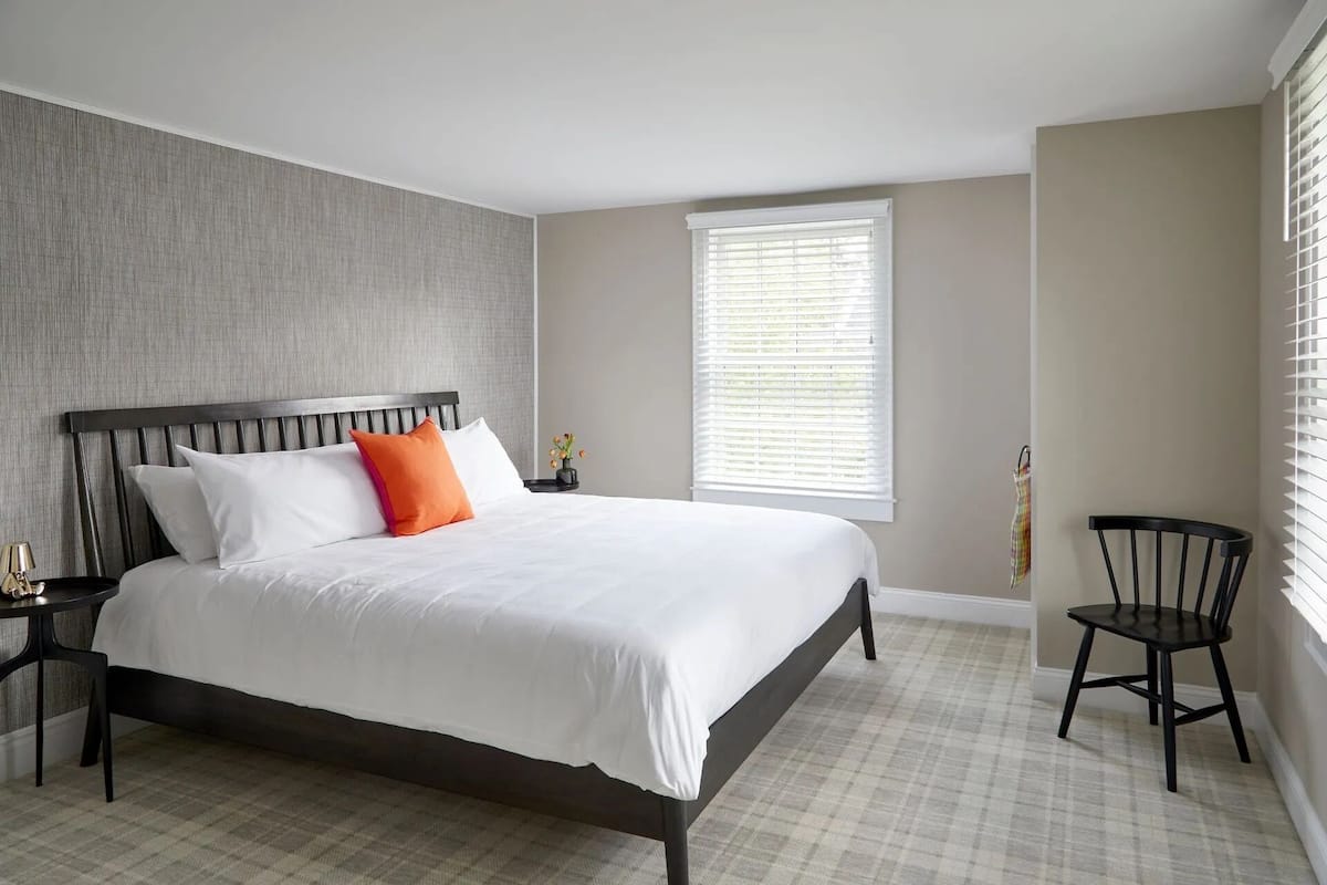 A neatly made bed with white and orange pillows, a dark wooden headboard, and a small table with a lamp beside it.